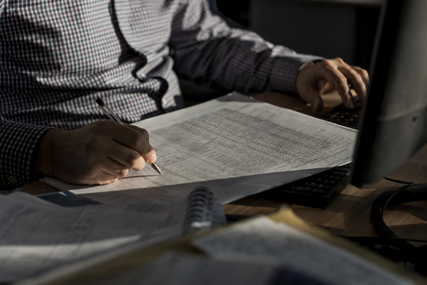 Hands at a desk, one on a keyboard and one annotating a printed report in pen.